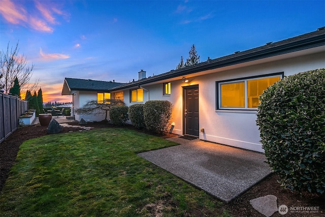 back of property at dusk with fence, a yard, a chimney, stucco siding, and a patio area