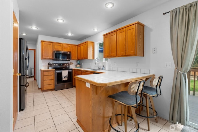 kitchen with tile countertops, a peninsula, light tile patterned flooring, a sink, and appliances with stainless steel finishes