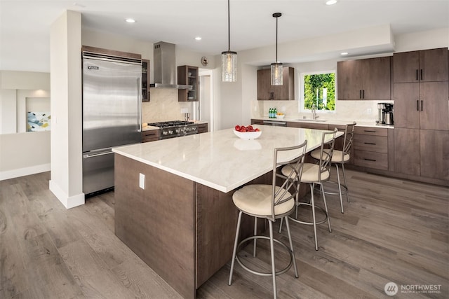 kitchen with a sink, a kitchen island, wood finished floors, appliances with stainless steel finishes, and wall chimney exhaust hood