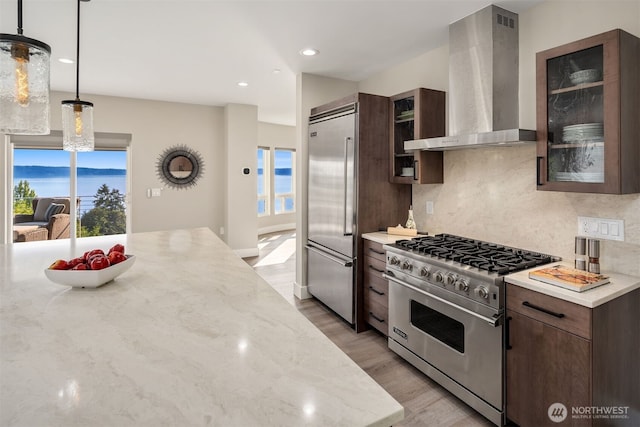 kitchen featuring decorative backsplash, dark brown cabinets, light wood-style floors, wall chimney exhaust hood, and premium appliances