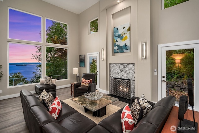 living area with a water view, a tiled fireplace, wood finished floors, a high ceiling, and baseboards