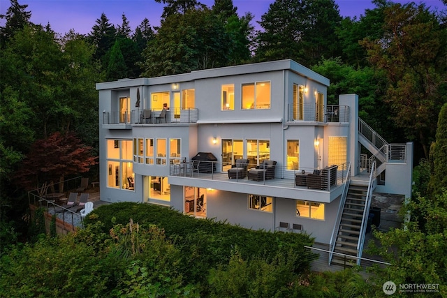 back of house at dusk with stucco siding, a balcony, and stairs