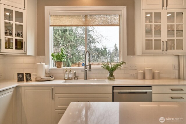 kitchen featuring a sink, decorative backsplash, light countertops, glass insert cabinets, and stainless steel dishwasher