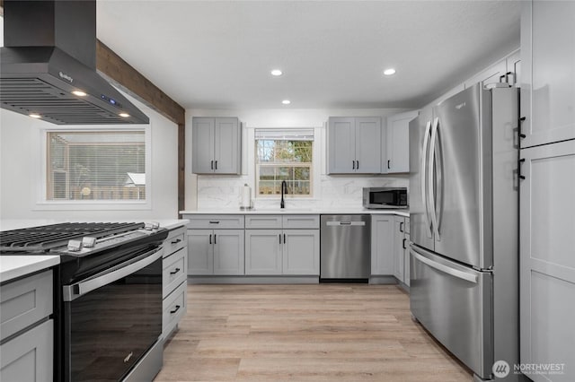 kitchen with light wood-type flooring, gray cabinets, a sink, stainless steel appliances, and wall chimney range hood