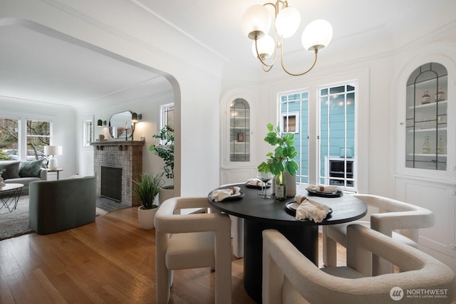 dining room featuring ornamental molding, wood finished floors, arched walkways, an inviting chandelier, and a brick fireplace