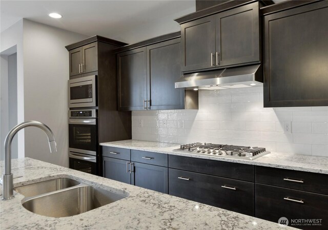 kitchen featuring light stone counters, a sink, appliances with stainless steel finishes, under cabinet range hood, and tasteful backsplash