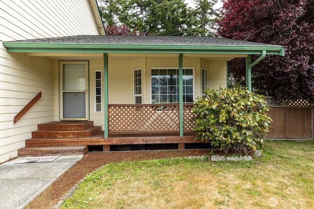 doorway to property featuring covered porch, a shingled roof, a lawn, and fence