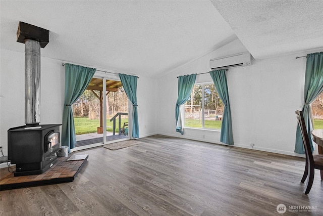 living area with a wall mounted AC, a textured ceiling, wood finished floors, a wood stove, and vaulted ceiling