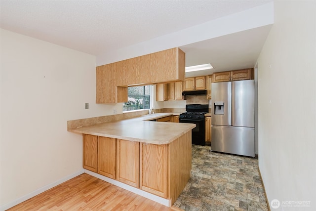 kitchen featuring under cabinet range hood, black gas range, light countertops, a peninsula, and stainless steel fridge