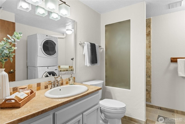 bathroom featuring visible vents, toilet, a textured wall, vanity, and stacked washer / drying machine