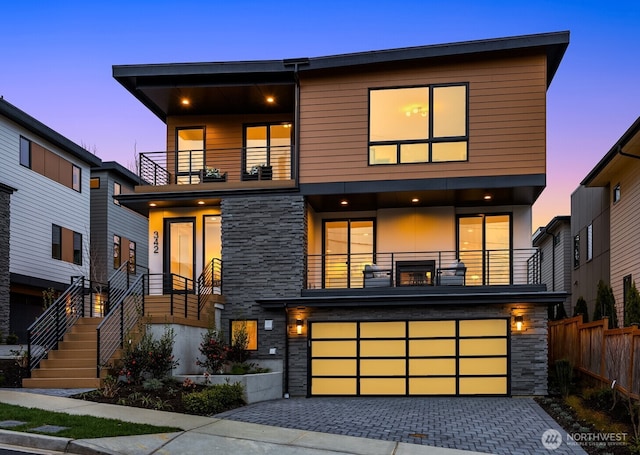 modern home featuring decorative driveway, stone siding, fence, a garage, and a balcony