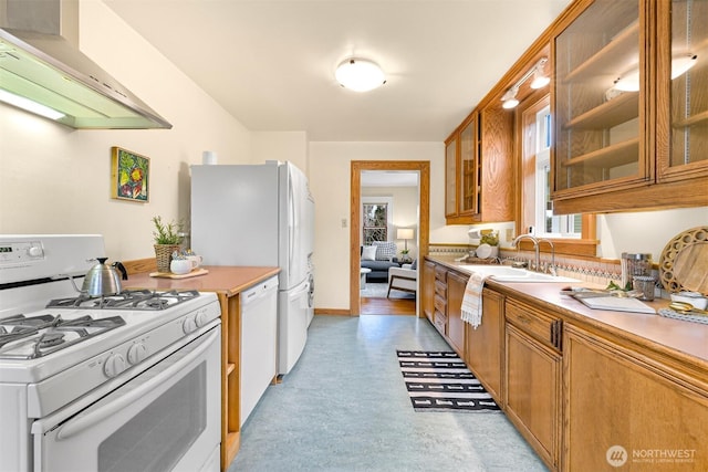 kitchen featuring white appliances, brown cabinetry, a sink, glass insert cabinets, and wall chimney range hood