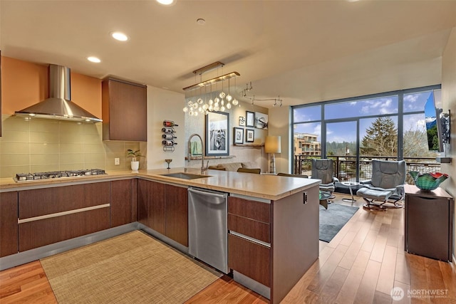 kitchen with a peninsula, stainless steel appliances, a sink, wall chimney exhaust hood, and open floor plan