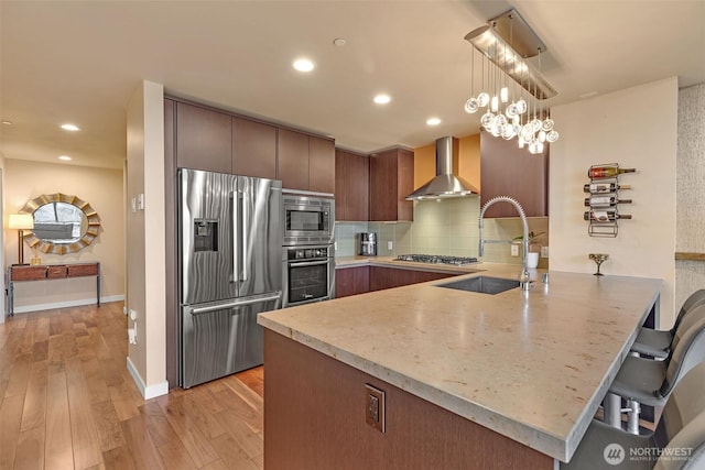 kitchen with a sink, backsplash, stainless steel appliances, a peninsula, and wall chimney exhaust hood