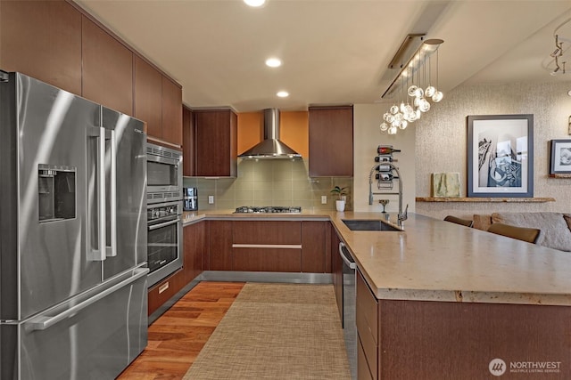 kitchen with a sink, a peninsula, appliances with stainless steel finishes, wall chimney exhaust hood, and light countertops