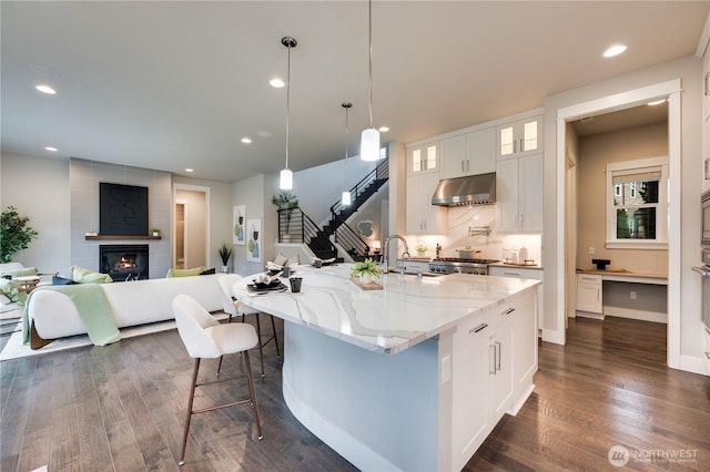 kitchen with light stone counters, dark wood finished floors, a sink, glass insert cabinets, and under cabinet range hood