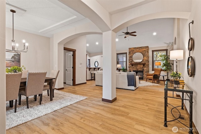 dining area with light wood-type flooring, ceiling fan with notable chandelier, arched walkways, a stone fireplace, and baseboards