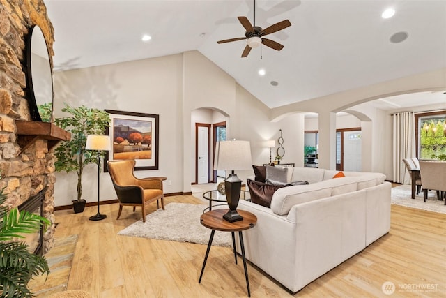 living area featuring a ceiling fan, wood finished floors, high vaulted ceiling, arched walkways, and a stone fireplace