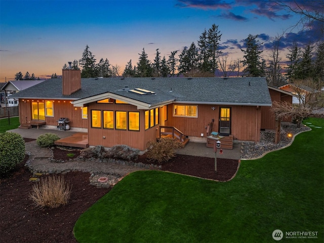 back of house at dusk with a shingled roof, a yard, a patio area, and a chimney