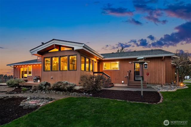 back of house at dusk with a lawn and a shingled roof