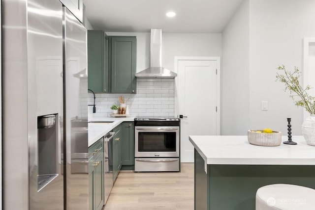 kitchen featuring wall chimney range hood, backsplash, stainless steel appliances, green cabinets, and light countertops