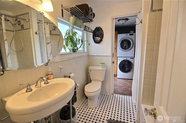 full bathroom featuring toilet, wainscoting, tiled shower, stacked washer / drying machine, and a sink