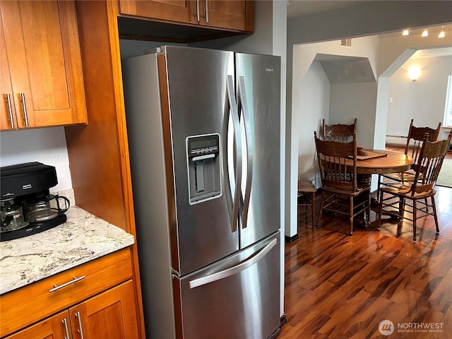 kitchen with light stone counters, brown cabinetry, stainless steel fridge with ice dispenser, arched walkways, and dark wood-type flooring