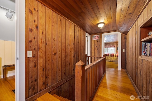 hallway with an upstairs landing, wood walls, wooden ceiling, and light wood-type flooring