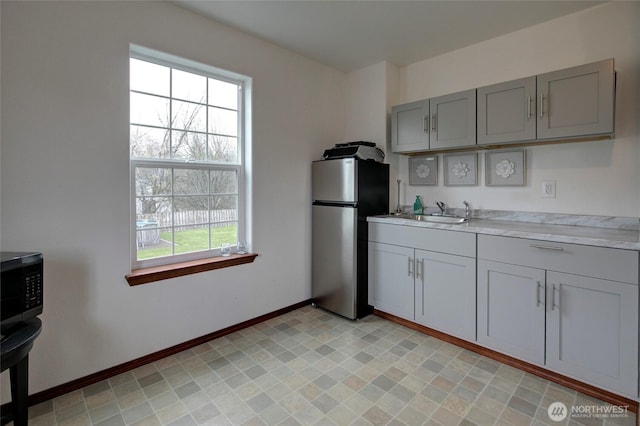 kitchen featuring baseboards, freestanding refrigerator, a sink, gray cabinetry, and light countertops