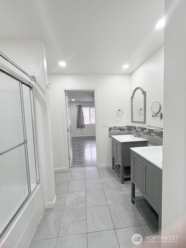 full bathroom featuring baseboards, shower / bath combination with glass door, two vanities, a sink, and backsplash