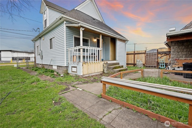 view of front of home with fence, roof with shingles, covered porch, a yard, and a garden