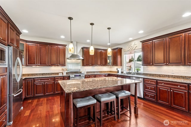 kitchen featuring a kitchen island, dark wood-style flooring, appliances with stainless steel finishes, wall chimney exhaust hood, and a sink