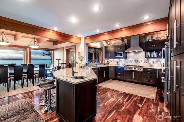 kitchen with a peninsula, wall chimney exhaust hood, light stone counters, a sink, and appliances with stainless steel finishes