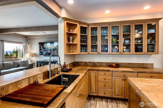 kitchen featuring a sink, open floor plan, glass insert cabinets, light wood-type flooring, and beamed ceiling