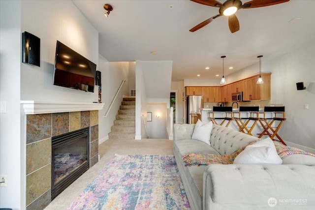 living area with baseboards, a tiled fireplace, stairway, light colored carpet, and a ceiling fan