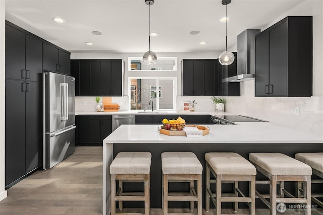 kitchen featuring decorative light fixtures, appliances with stainless steel finishes, a peninsula, wall chimney exhaust hood, and dark cabinets