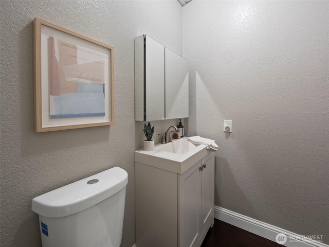 half bathroom with baseboards, toilet, vanity, and a textured wall