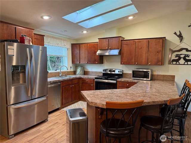 kitchen featuring under cabinet range hood, a sink, vaulted ceiling with skylight, appliances with stainless steel finishes, and a peninsula