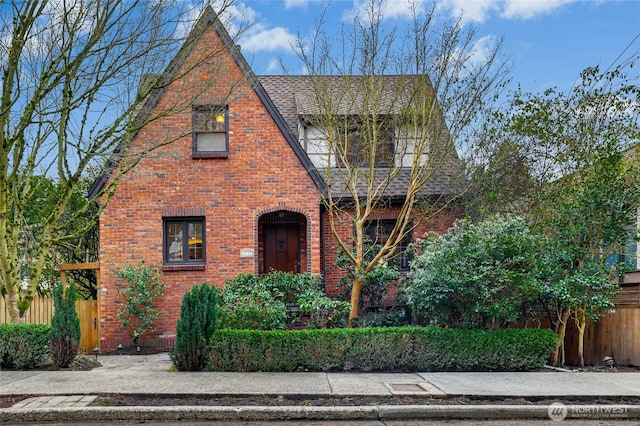 english style home with fence, brick siding, and a shingled roof