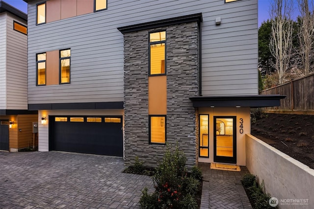 modern home featuring a garage, decorative driveway, fence, and stone siding