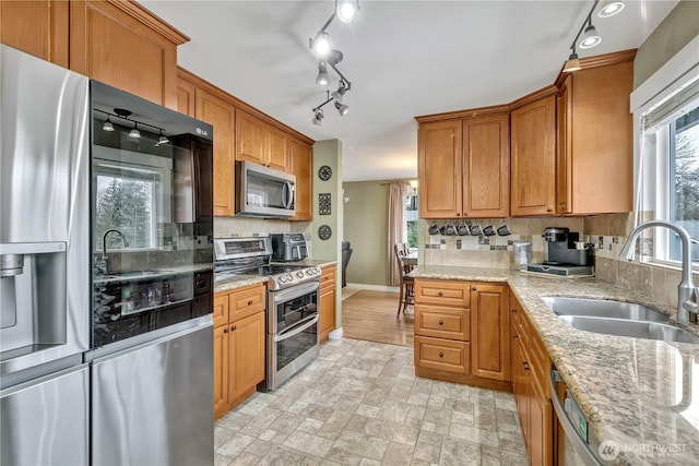 kitchen with light stone countertops, brown cabinetry, a sink, stainless steel appliances, and backsplash