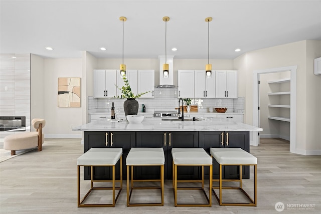 kitchen featuring a kitchen bar, wall chimney range hood, an island with sink, and white cabinets