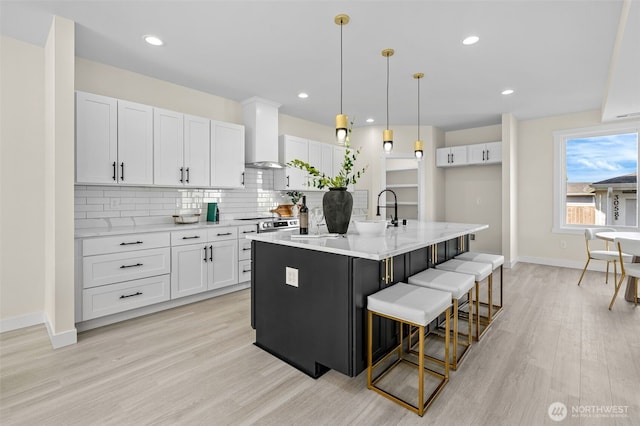 kitchen featuring tasteful backsplash, wall chimney range hood, light wood-style flooring, white cabinets, and a kitchen island with sink