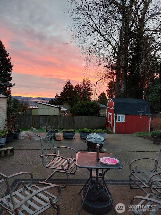 patio terrace at dusk featuring outdoor dining space, a storage shed, fence, and an outbuilding