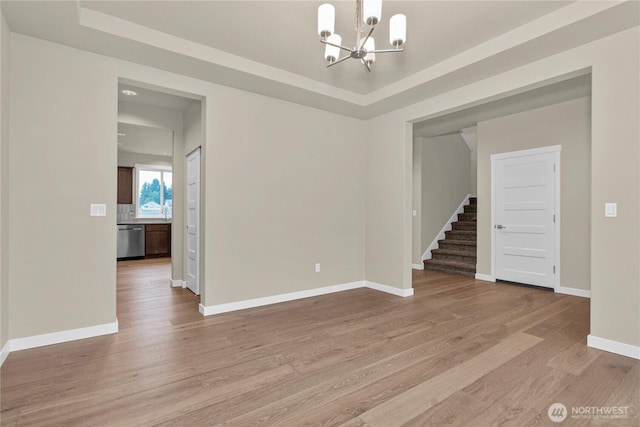 empty room featuring light wood-type flooring, a raised ceiling, a chandelier, and stairs