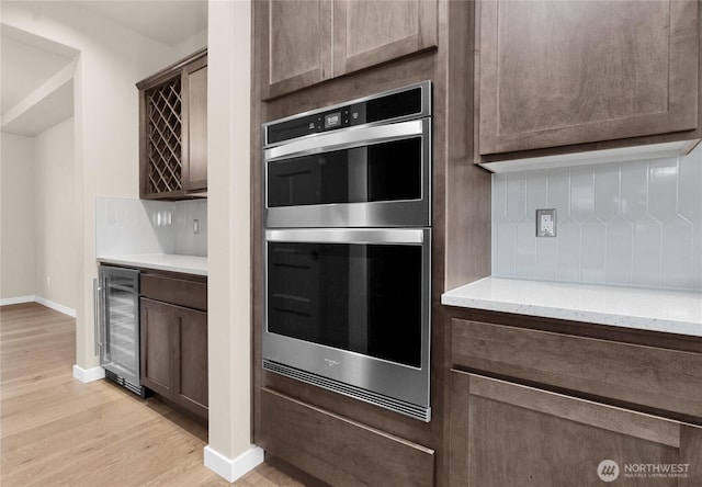 kitchen with beverage cooler, light wood finished floors, dark brown cabinets, double oven, and tasteful backsplash