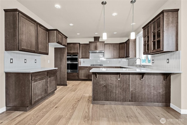 kitchen featuring a peninsula, cooktop, dark brown cabinets, light wood-style floors, and double oven