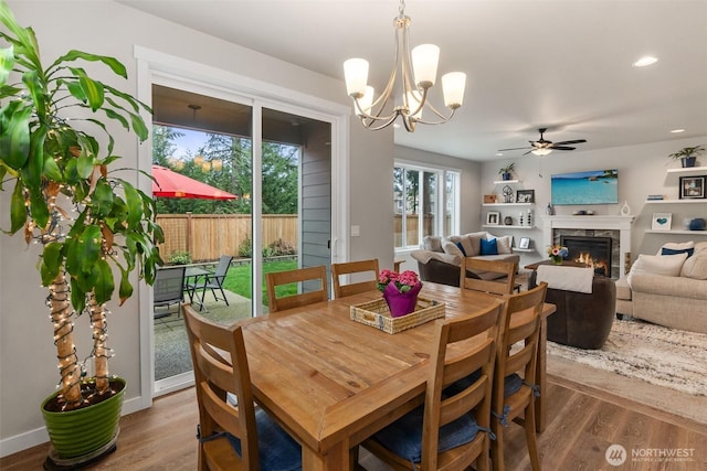 dining area featuring baseboards, recessed lighting, ceiling fan with notable chandelier, a warm lit fireplace, and wood finished floors