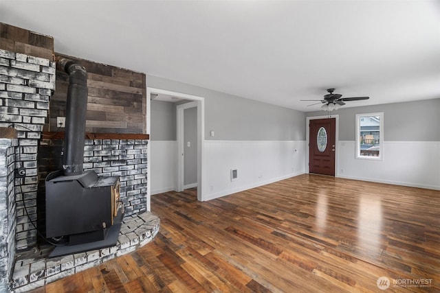 unfurnished living room featuring visible vents, wainscoting, a wood stove, wood finished floors, and a ceiling fan