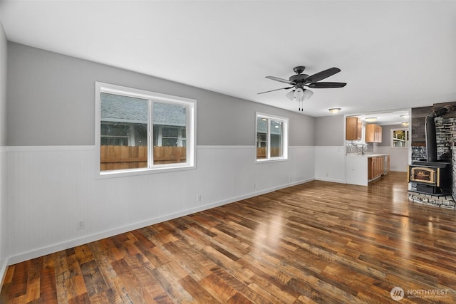 unfurnished living room featuring a wainscoted wall, a ceiling fan, a sink, dark wood finished floors, and a wood stove